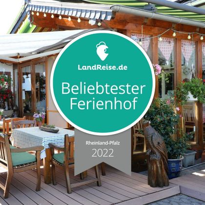 Eine einladende Terrasse mit Holzmöbeln und vielen Pflanzen. Das Bild zeigt ein Zertifikat für den "beliebtesten Ferienhof" in Rheinland-Pfalz 2022.