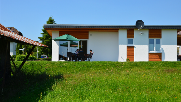 Ein modernes Haus mit einer Terrasse und einem grünen Rasen. Auf der Terrasse sitzen Personen unter einem Sonnenschirm.