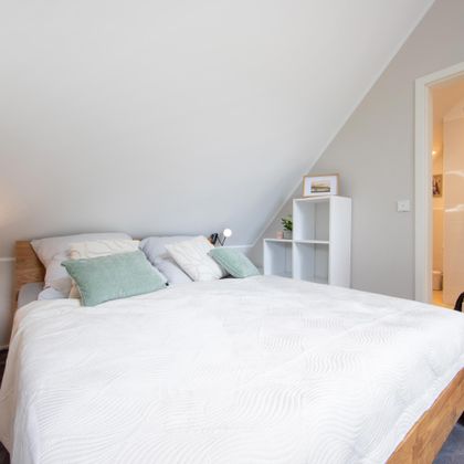Ein helles Schlafzimmer mit einem großen Bett und gemütlichen Kissen. Im Hintergrund ist ein modernes Bad mit Dusche sichtbar.
