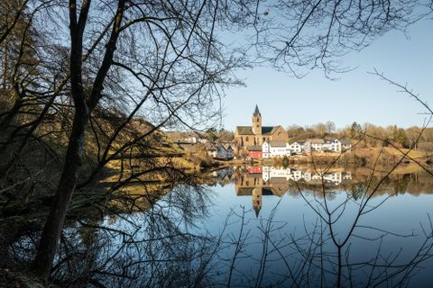 A peaceful landscape with a clear lake that reflects the church and the surrounding buildings. Trees and a clear sky can be seen in the background.