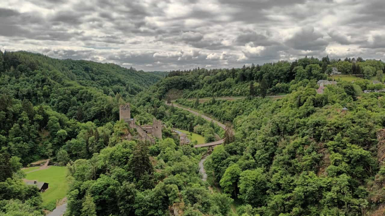 Eine beeindruckende Landschaft mit grünen Hügeln und einer alten Burgruine. Der Himmel ist bewölkt und die Umgebung ist von Bäumen umgeben.
