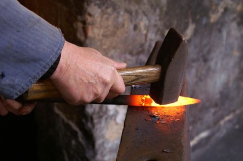 Ein Schmied bearbeitet ein glühendes Metallstück mit einem Hammer auf einem Amboss.