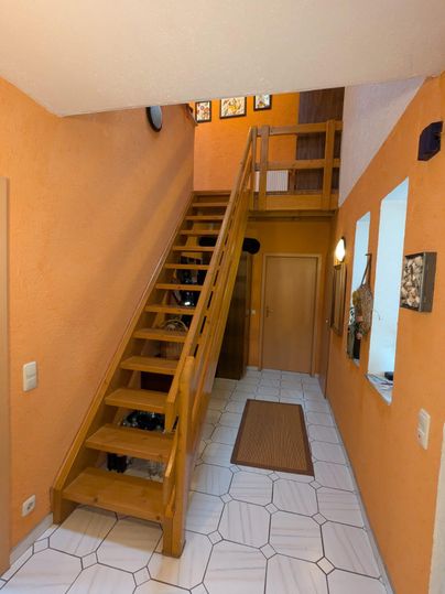 Ein heller Flur mit orangefarbenen Wänden und einer Holztreppe. Der Boden ist mit weißen Fliesen und einem Teppich in der Mitte gestaltet.