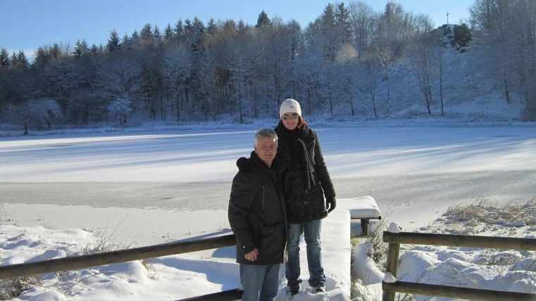 Ein verschneites Landschaftsbild mit einem gefrorenen See. Zwei Personen stehen auf einer Holzplanke vor dem See.