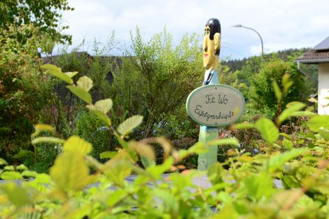 Ein bunter Holzschild mit einer Figur steht zwischen Pflanzen. Darauf steht der Schriftzug „Ich bin Eifgenhüpf!“