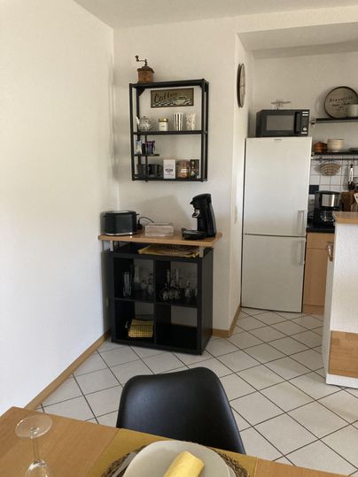 Eine moderne Küche mit einem kleinen Tisch und schwarzen Stühlen. Im Hintergrund befinden sich ein Kühlschrank, eine Mikrowelle und eine Kaffeekanne auf einem schwarzen Regal.