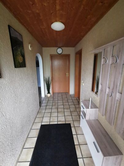 Ein heller Flur mit einer Holzbalkendecke und gefliesten Böden. An der Wand befindet sich eine Garderobe und ein Spiegel.