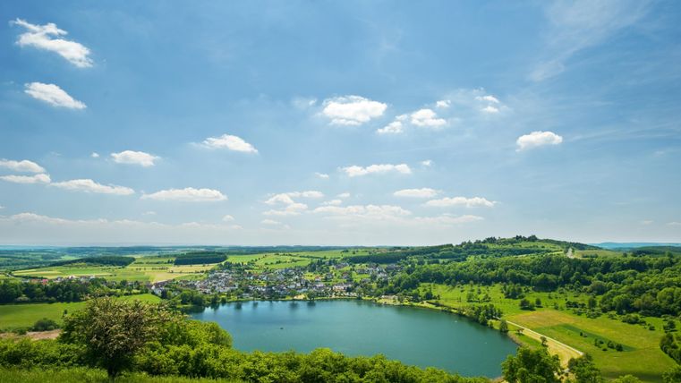 Eine malerische Landschaft mit einem ruhigen See und üppigen grünen Wiesen. Der Himmel ist blau mit einigen weißen Wolken.