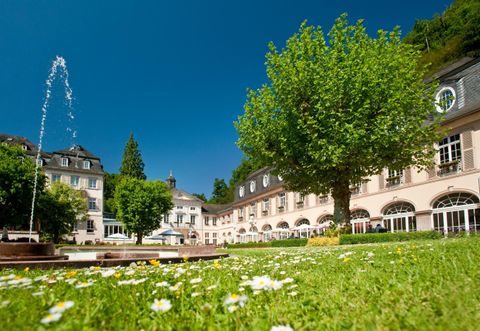 Ein schöner Garten mit einem großen Baum und einer Wasserfontäne. Im Hintergrund befindet sich ein elegantes Gebäude unter einem klaren blauen Himmel.