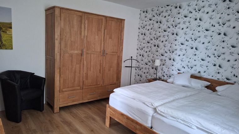 Ein schlichtes Schlafzimmer mit einem Doppelbett und einem großen Kleiderschrank aus Holz. Neben dem Bett steht ein schwarzer Sessel und eine Wand mit floralem Muster.