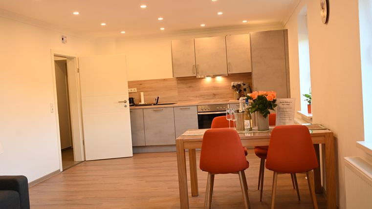 Ein modernes, helles Küchen- und Esszimmer mit Holzfußboden. Der Tisch ist mit Blumen dekoriert und es gibt orangefarbene Stühle.