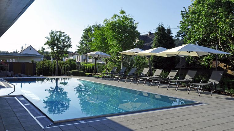 Ein klarer Pool umgeben von Liegen und Sonnenschirmen. Die Umgebung ist grün mit Bäumen und einem hellen Himmel.