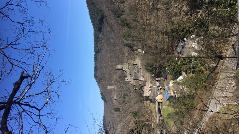 Eine alte Ruine in einer hügeligen Landschaft unter einem klaren blauen Himmel. Im Vordergrund sind kahle Bäume sichtbar.