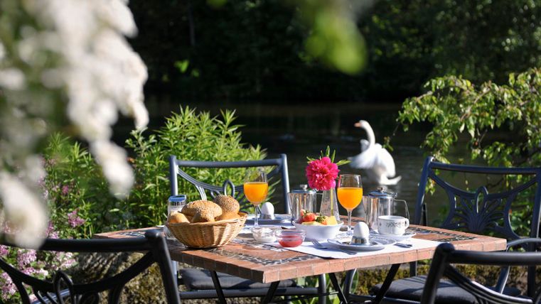 Ein einladender Tisch im Freien, gedeckt mit Frühstück und frischen Brötchen. Im Hintergrund sind Blumen und eine ruhige Wasseroberfläche zu sehen.