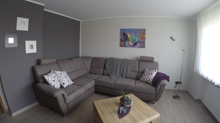 Ein modernes Wohnzimmer mit einer beigen L-förmigen Couch und dekorativen Kissen. Der Raum verfügt über einen Holztisch und einen eleganten Stehlampe, ergänzt durch ein farbenfrohes Bild an der Wand.