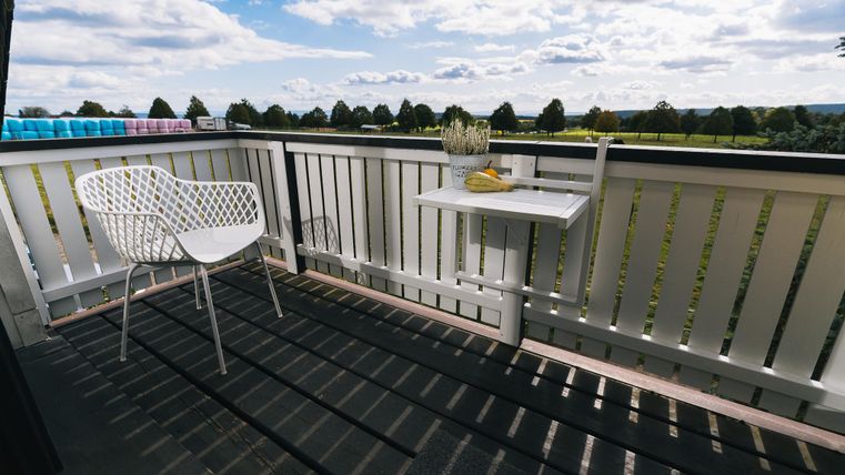 Ein gemütlicher Balkon mit einem weißen Stuhl und einem kleinen Tisch. Im Hintergrund erstreckt sich eine grüne Landschaft unter einem teilweise bewölkten Himmel.