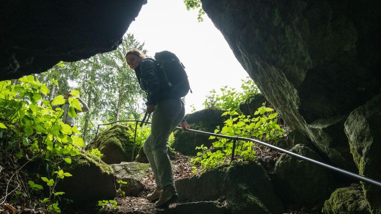 Zwei Personen steigen aus einer Höhle ins Freie. Umgeben von Felsen und Pflanzen, führt ein Geländer den Weg hinauf.