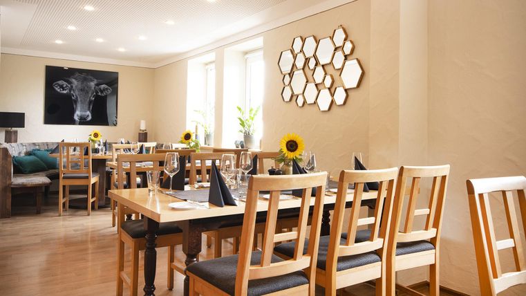 Ein modernes Restaurant mit holzernen Tischen und Stühlen. Die Wände sind hell gestrichen und mit einem Kunstwerk sowie Spiegeln dekoriert.