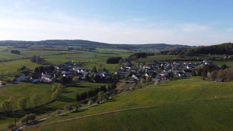 Eine malerische Landschaft mit sanften Hügeln und einem kleinen Dorf. Das Dorf ist von grünen Feldern umgeben und liegt unter einem klaren blauen Himmel.