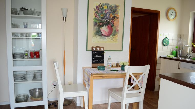 Ein gemütlicher Essbereich mit einem kleinen Tisch und zwei Stühlen. An der Wand hängt ein buntes Blumenbild, und in der Ecke steht ein Glasregal.