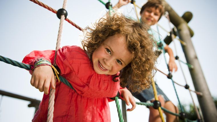 Zwei Kinder spielen auf einem Spielplatz. Ein Mädchen mit lockigem Haar lächelt fröhlich, während ein Junge im Hintergrund klettert.