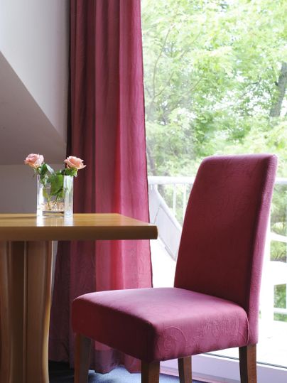 Ein eleganter Raum mit einem roten Stuhl und einem kleinen Tisch. Durch das Fenster fällt Licht auf die frischen Blumen in der Vase.