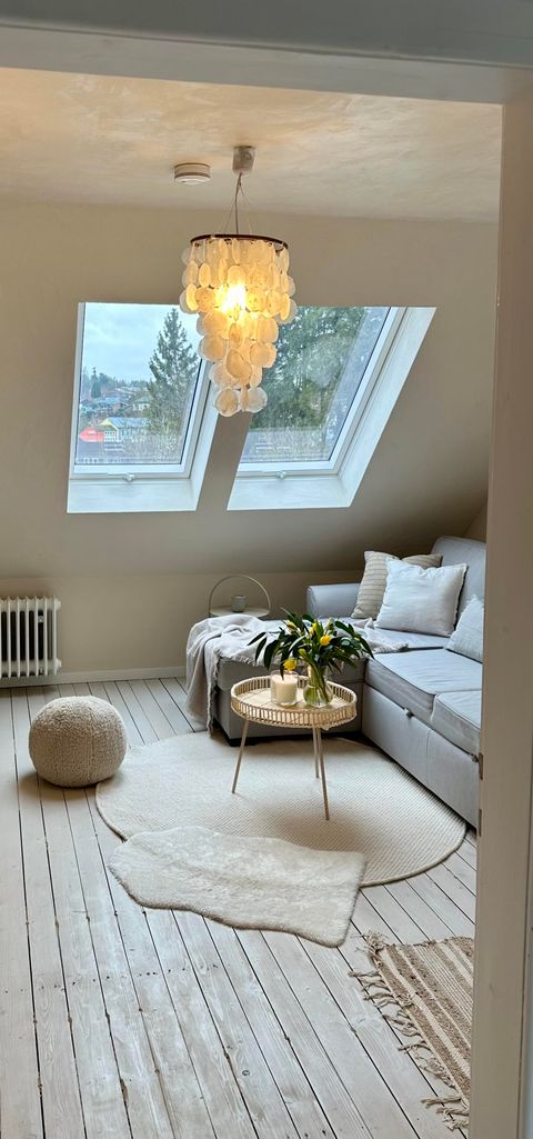Ein gemütlicher Raum mit einer hellen Couch und großen Fenstern. Das Lichtspiel von der Deckenlampe schafft eine einladende Atmosphäre.