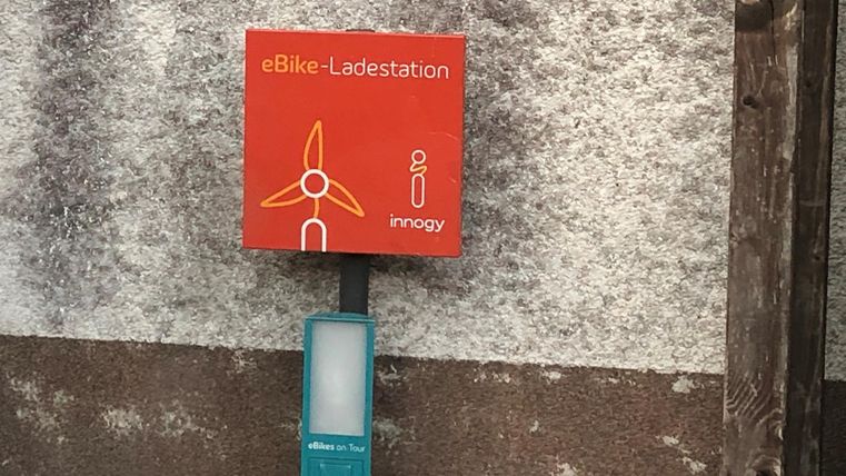 Eine eBike-Ladestation mit einem orangefarbenen Schild. Sie befindet sich an einer grauen Wand.