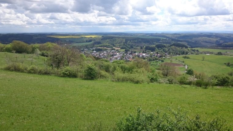 Eine malerische Aussicht auf ein kleines Dorf im Tal, umgeben von grünen Wiesen und Hügeln. Der Himmel ist bewölkt und die Landschaft wirkt beruhigend.