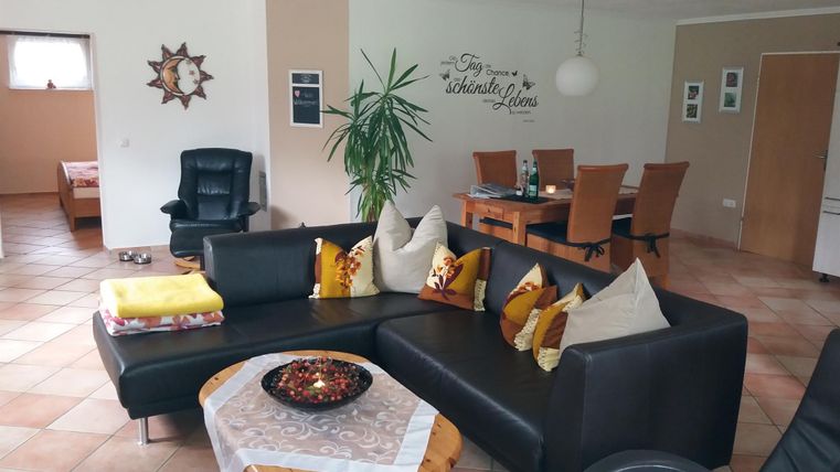 Ein gemütliches Wohnzimmer mit einer großen Sofa-Ecke und dekorativen Kissen. Im Hintergrund sind ein Esstisch und eine Pflanze zu sehen.