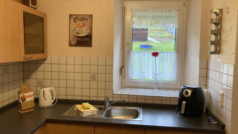 Eine helle Küche mit Holzschränken und einem großen Fenster. Auf der Arbeitsplatte stehen eine Kaffeekanne und einige Kochutensilien.