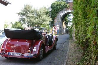 Ein rotes Oldtimer-Cabriolet steht an einer gepflasterten Straße. Im Hintergrund ist ein Tor mit grüner Vegetation zu sehen.