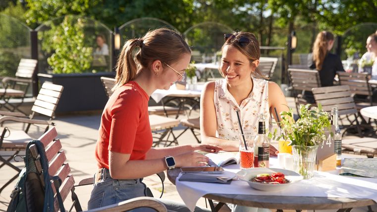 Zwei Frauen sitzen fröhlich an einem Tisch im Freien und genießen ihre Zeit zusammen. Auf dem Tisch stehen Getränke und ein kleiner Teller mit Essen.