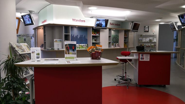Ein modernes Informationszentrum mit mehreren Theken und TV-Bildschirmen. Die Räume sind hell und freundlich gestaltet, mit Pflanzen im Hintergrund.