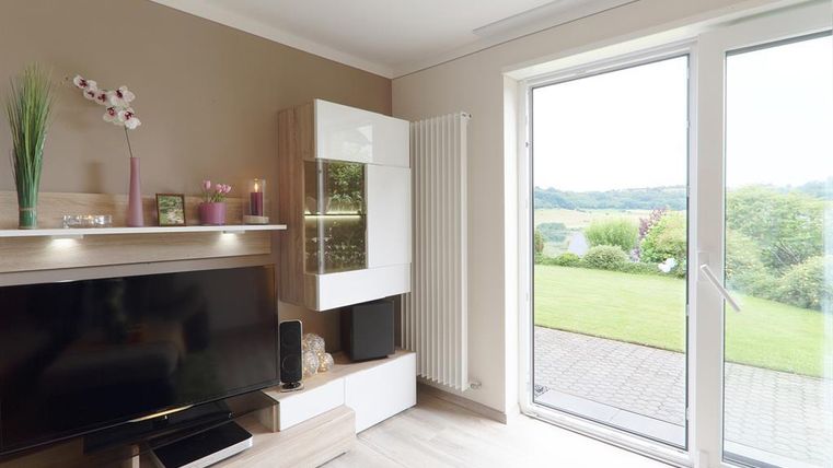 Ein modernes Wohnzimmer mit einem Fernseher und einer großen Fensterfront. Durch das Fenster ist ein grüner Garten und die Landschaft sichtbar.