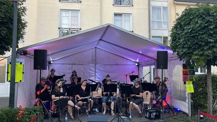A band is playing under a tent roof in an urban setting. In the background, trees and a building can be seen.