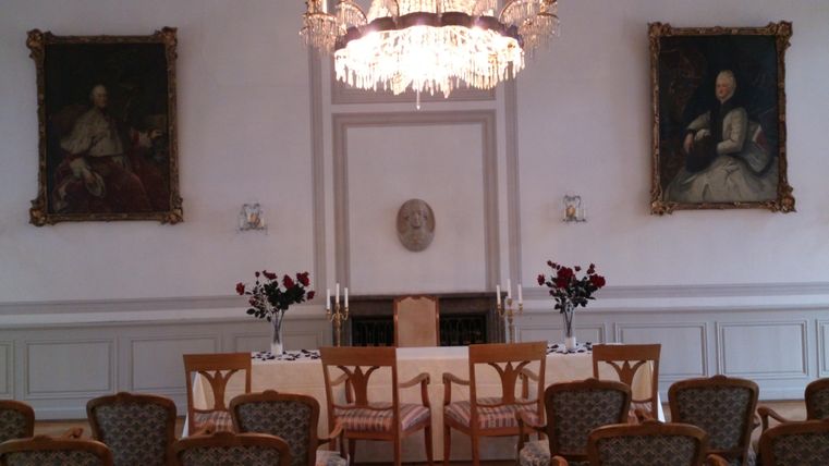 Ein eleganter Raum mit einem Kronleuchter und zwei großen Gemälden an den Wänden. Ein gedeckter Tisch mit Stühlen steht in der Mitte, umgeben von dekorativen Blumen.