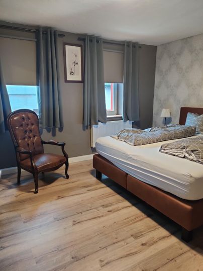 Ein gemütliches Schlafzimmer mit einem Doppelbett und einem Sessel. Die Wände sind hell dekoriert, und das Licht strömt durch die Fenster.