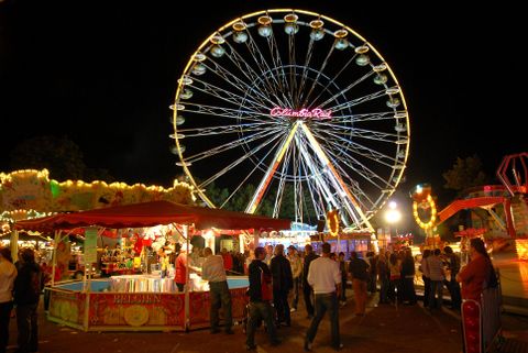 Ein Riesenrad leuchtet in der Nacht über einem Jahrmarkt. Menschen schlendern zwischen Ständen und Fahrgeschäften.