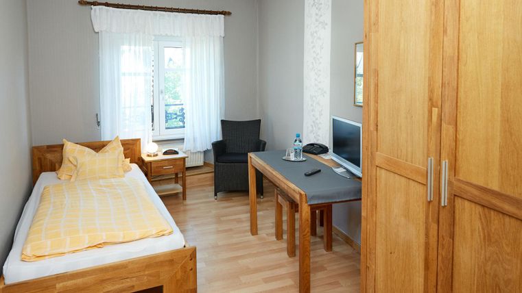 Ein einfaches Zimmer mit einem Einzelbett, einem Tisch und einem Stuhl. Die Wände sind hell und es gibt große Fenster, die für viel Licht sorgen.