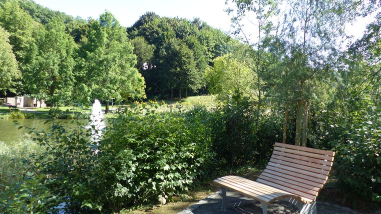 Eine ruhige Parklandschaft mit einem kleinen See und einem Springbrunnen. Eine Holzliege steht am Ufer, umgeben von grünen Büschen und Bäumen.