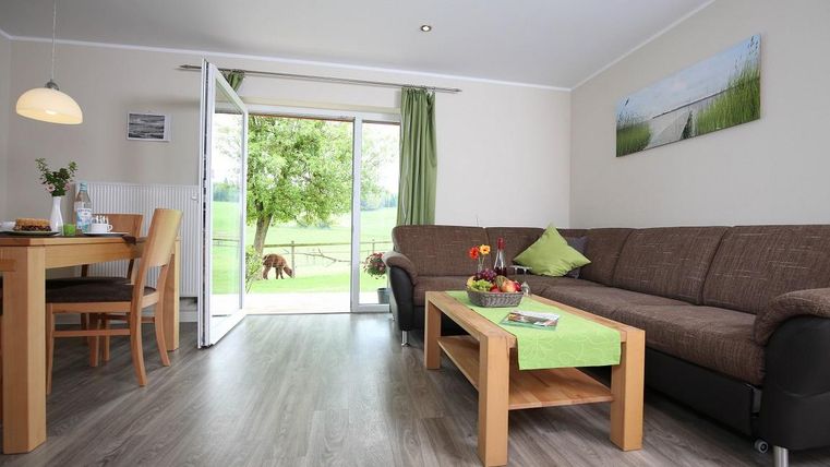 Ein helles Wohnzimmer mit einer gemütlichen Couch und einem Tisch. Große Fenster bieten einen Blick ins Freie auf eine grüne Wiese.