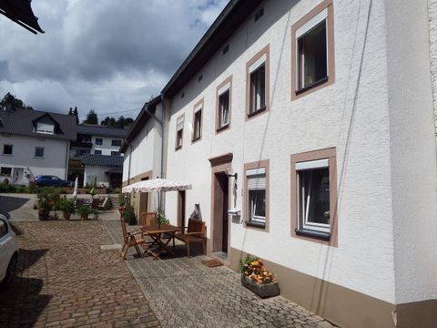 Ein helles, zweigeschossiges Haus mit weißen Wänden und braunen Fensterrahmen. Vor dem Haus steht ein Tisch mit Stühlen und einige Blumen sind zu sehen.