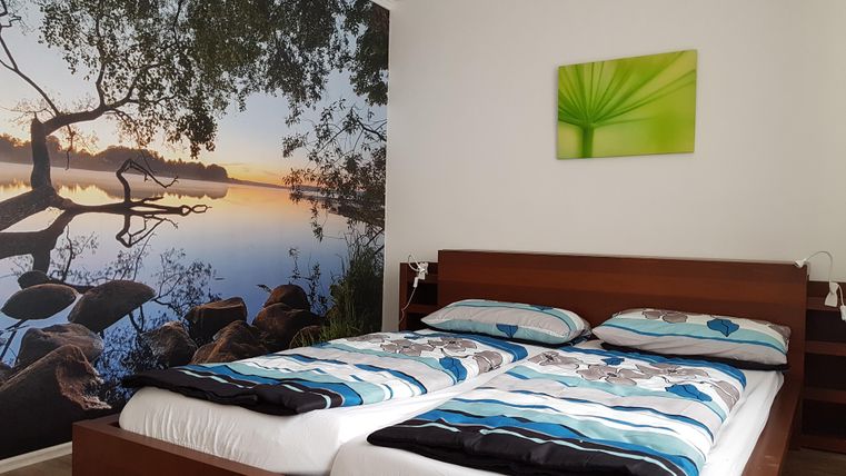 Ein gemütliches Schlafzimmer mit einem Doppelbett und einem großen Wandbild eines ruhigen Sees. An der Wand hängt ein grünes Bild mit einer Pflanze.