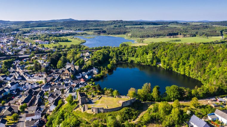 Eine malerische Landschaft mit einem ruhigen See, umgeben von grünen Wäldern und sanften Hügeln. Im Vordergrund sind kleine Häuser und Ruinen sichtbar.