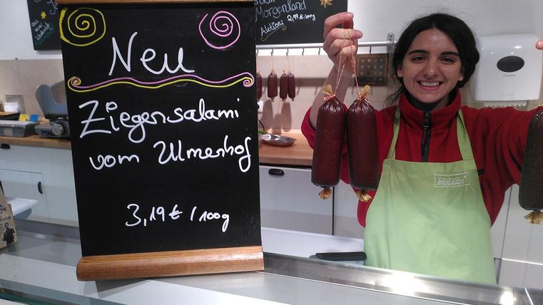 Eine Frau steht hinter einem Ladentisch und hält zwei Wurstwaren in der Hand. Neben ihr steht ein Schild mit der Aufschrift „Neu: Ziegensalami vom Ulmenhof“.