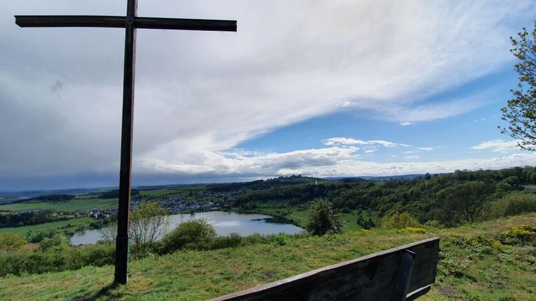 Ein Holzsteg führt über einen ruhigen See, umgeben von grünem Gras und Bäumen. Der Himmel ist klar mit einigen Wolken.