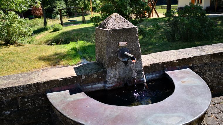 Ein Brunnen im Dauener Kurpark mit fließendem Wasser, umgeben von Bäumen und einem Spielplatz im Hintergrund.