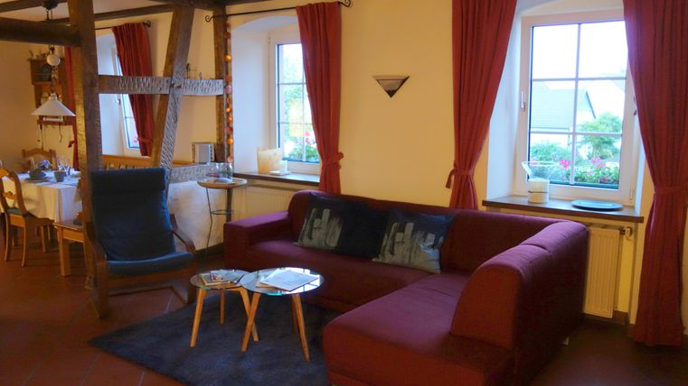 Ein gemütliches Wohnzimmer mit einem roten Sofa und einem schwarzen Teppich. Große Fenster mit roten Vorhängen sorgen für viel Tageslicht.
