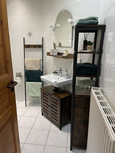 Ein modernes Badezimmer mit weißen Wänden und einem Waschbecken. Handtücher und Pflegeprodukte sind ordentlich auf Regalen und einem Handtuchhalter angeordnet.